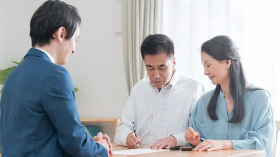スーツ姿の担当者と向かい合い、書類にサインをする中年夫婦の様子を捉えた場面